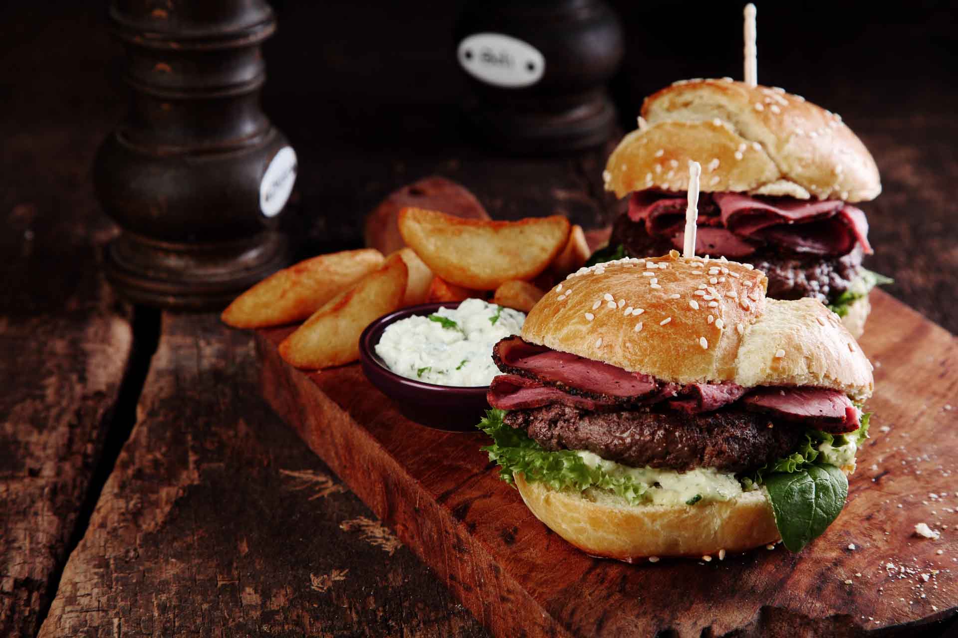 Leckere Gourmet-Steak-Burger mit Schinkenscheiben auf einem Holztablett mit Kartoffelspalten und Dip-Soße.