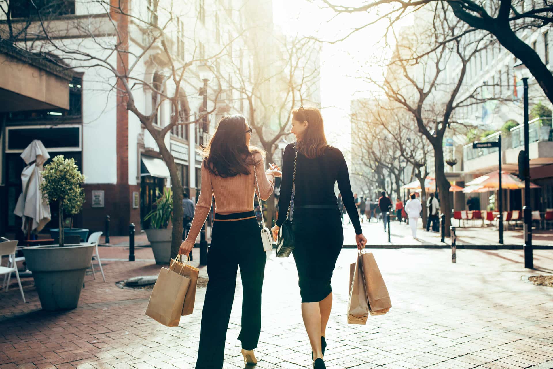 Rückansicht von zwei Freundinnen, die mit Einkaufstaschen auf der Stadtstraße spazieren gehen. Weibliche Shopper tragen Einkaufstaschen, während sie die Straße entlang gehen.