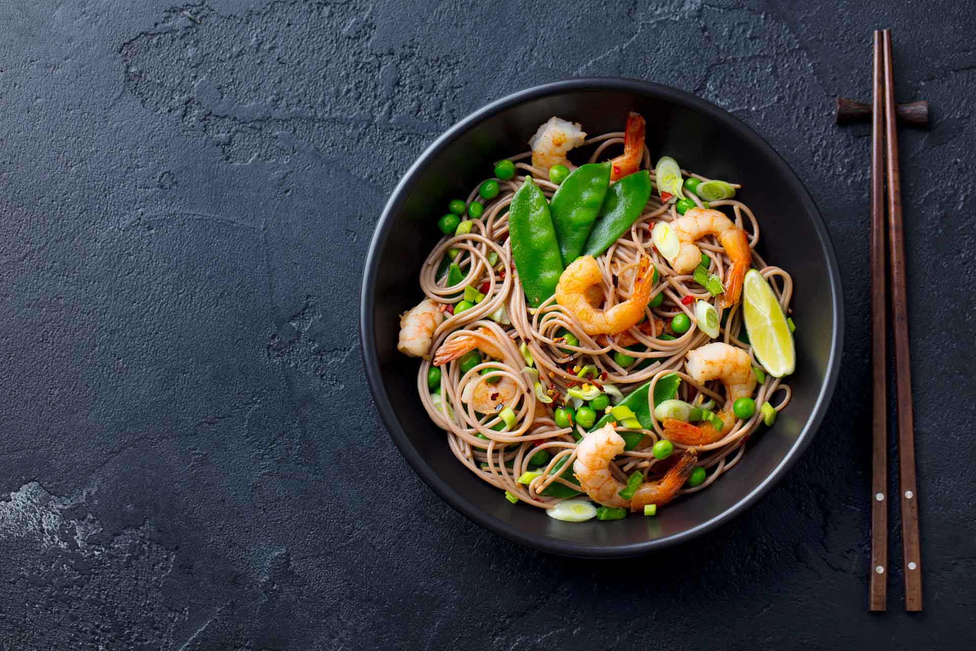 Stir fry noodles with vegetables and shrimps in black bowl. Slate background.