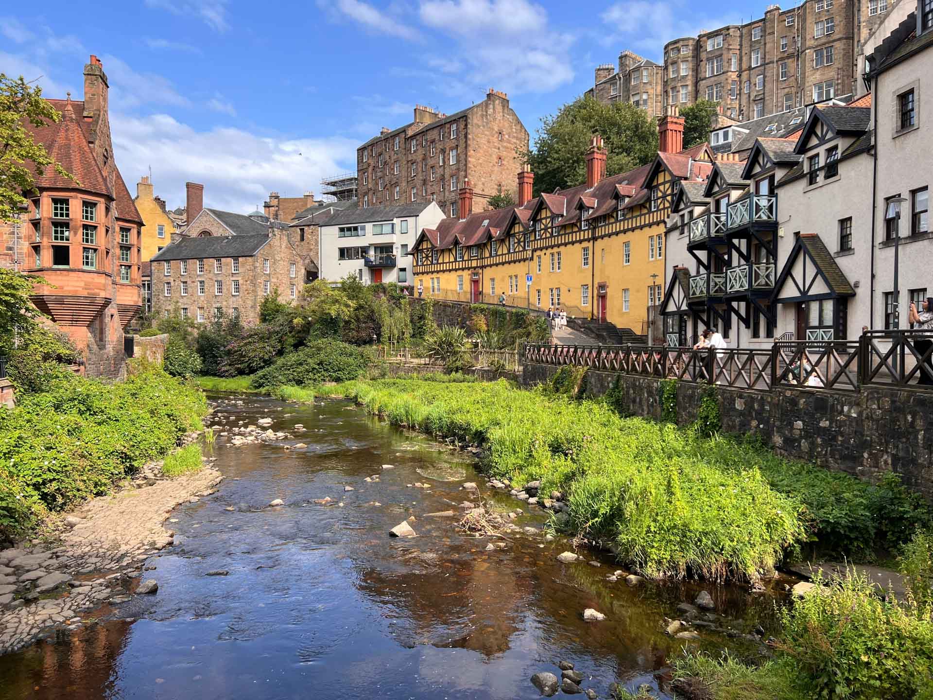 Scène paisible du village de Dean, à Édimbourg, avec un ruisseau qui coule dans une végétation luxuriante. Des bâtiments historiques et colorés bordent la rive, mêlant architecture médiévale et moderne sous un ciel radieux.
