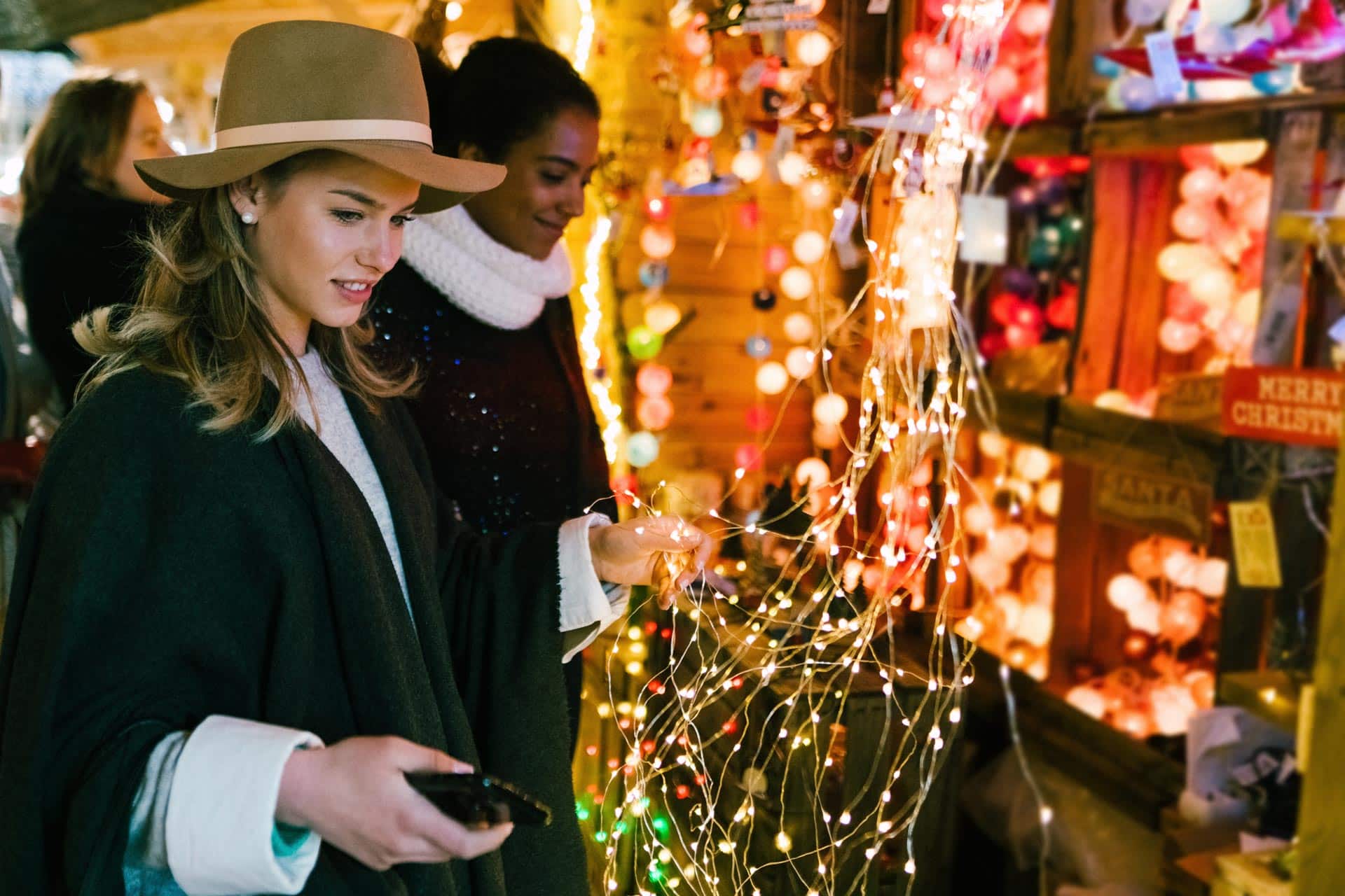 Two women windowshopping at festive market with string lights and ornaments.